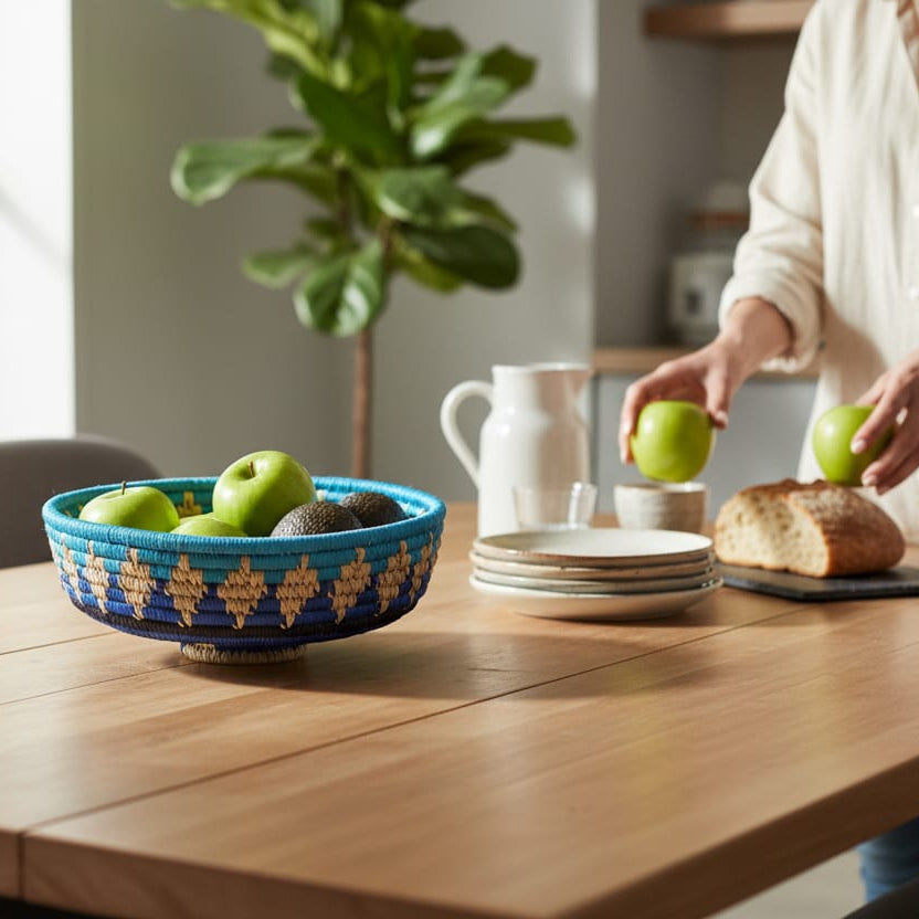 Person arranging apples around a blue bowl on a wooden table with a plant in the background.